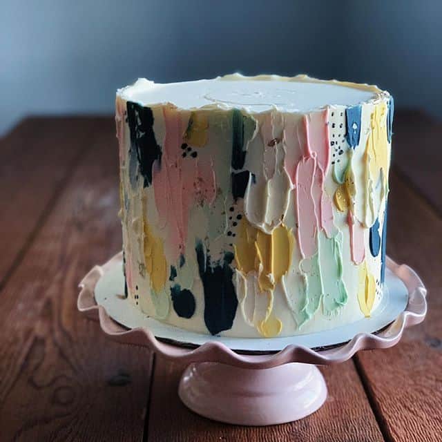 A hand painted wedding cake sits on a pink cake stand atop a rustic wooden table